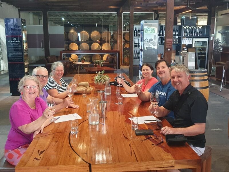 Group of six guests enjoying a wine tasting at a Coonawarra cellar door, seated around a long timber table.