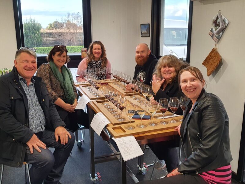 A group of seven people seated around a table, smiling and enjoying a wine tasting session indoors with a variety of wine glasses in front of them.