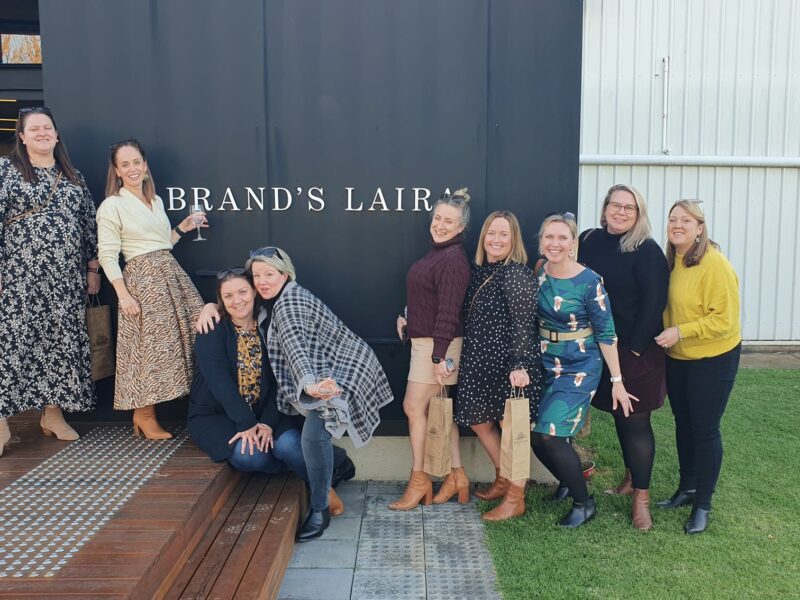 A group of nine women posing and smiling outside the entrance of Brand's Laira winery, enjoying their visit on a sunny day.