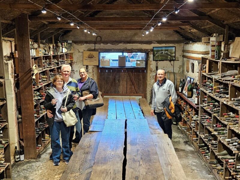 A group of four people standing inside a historic wine cellar, surrounded by shelves filled with old wine bottles, smiling for the camera.