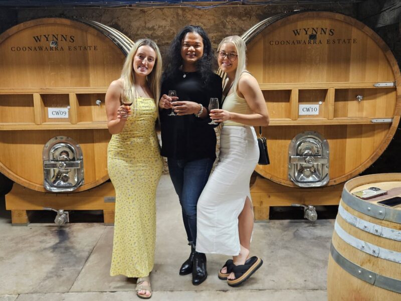 Three women smiling and holding glasses of wine while posing in front of large wooden wine barrels at Wynns Coonawarra Estate.