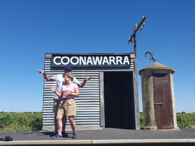 A couple posing and pointing in front of a corrugated iron building with a "Coonawarra" sign on a clear sunny day.