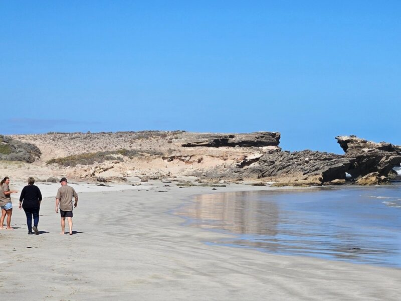 Three people walking barefoot along a sandy beach with rugged rock formations and blue skies in the background.