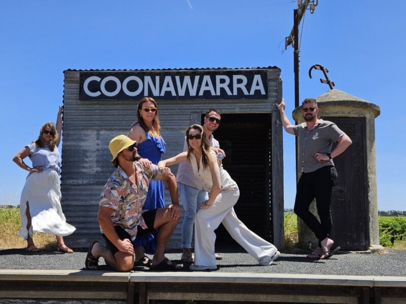 A group of six people striking fun poses at the historic Coonawarra Railway Siding, set against a backdrop of vineyards and a clear blue sky.