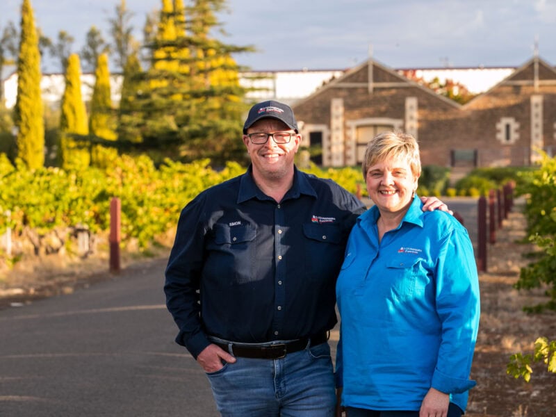 Couple smiling in a vineyard with historic winery building in the background, bathed in golden light.