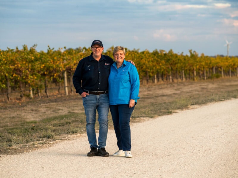 Couple standing on a vineyard road during golden hour, smiling at the camera.