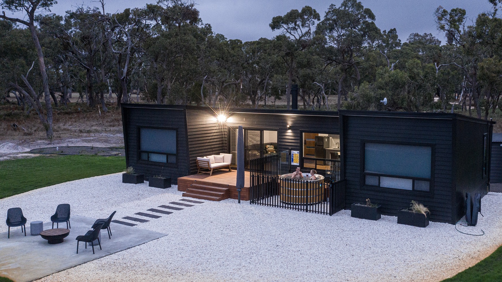 Modern black eco-retreat with cedar hot tub, deck seating, and firepit area, nestled in native bushland at dusk.